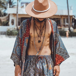 May include: A man wearing a brown fedora with a white band, a blue and red patterned kimono, and matching patterned shorts. He is wearing multiple necklaces with beads and charms.