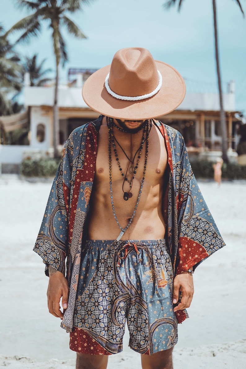 May include: A man wearing a brown fedora with a white band, a blue and red patterned kimono, and matching patterned shorts. He is wearing multiple necklaces with beads and charms.