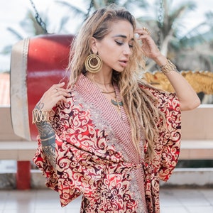 Puede incluir: Una mujer con un kimono estampado en rojo y dorado con un collar de oro y grandes pendientes de aro dorados. Tiene el pelo largo y rubio con rastas.
