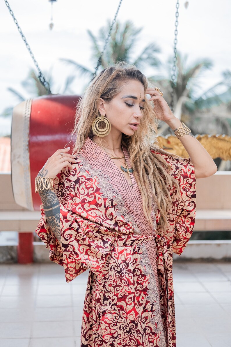 Puede incluir: Una mujer con un kimono estampado en rojo y dorado con un collar de oro y grandes pendientes de aro dorados. Tiene el pelo largo y rubio con rastas.