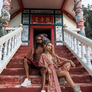 Puede incluir: Una pareja vestida con t&uacute;nicas rojas y doradas posa en los escalones de un templo chino tradicional. El templo tiene un techo rojo y paredes blancas con intrincados tallados y pinturas. La pareja est&aacute; sentada en los escalones, la mujer a la derecha y el hombre a la izquierda. La mujer tiene el pelo largo y rubio y lleva una t&uacute;nica roja y dorada con una larga cola. El hombre tiene el pelo largo y negro y lleva una t&uacute;nica roja y dorada con una cola corta. El templo est&aacute; decorado con s&iacute;mbolos y caracteres chinos tradicionales. El texto en el templo dice "高音 韻".