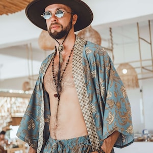 May include: A person wearing a blue and gold patterned kimono with a brown leather cord necklace and a brown hat. The person is wearing sunglasses and has a beard.