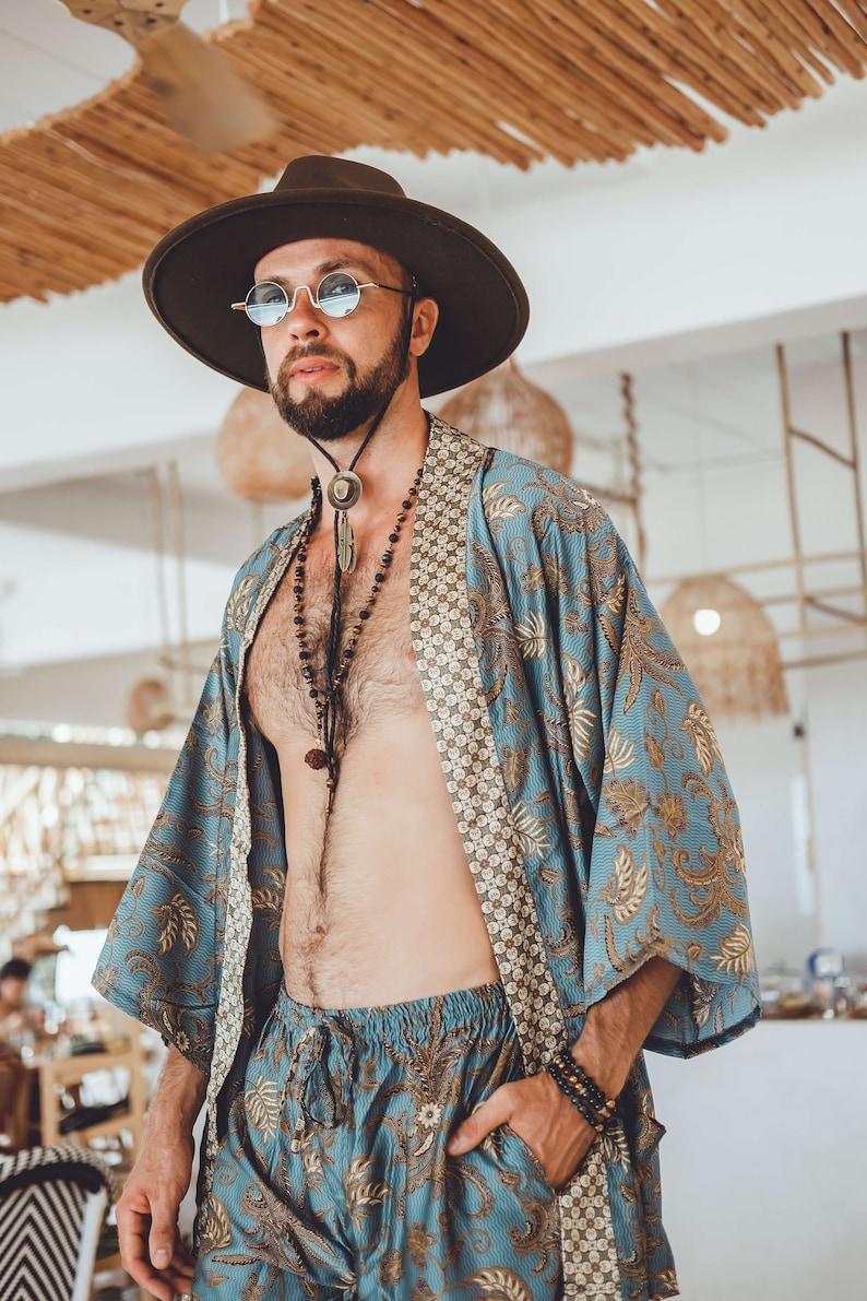 May include: A person wearing a blue and gold patterned kimono with a brown leather cord necklace and a brown hat. The person is wearing sunglasses and has a beard.