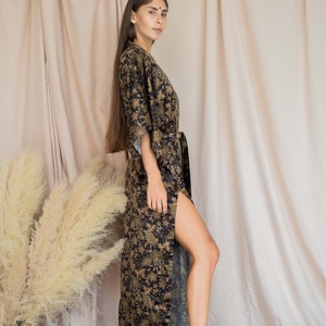 May include: A long, black and gold floral print kimono-style robe with a matching belt. The robe has a high slit on the side and is paired with black ankle boots. The background features neutral-toned fabric and dried ornamental grass.