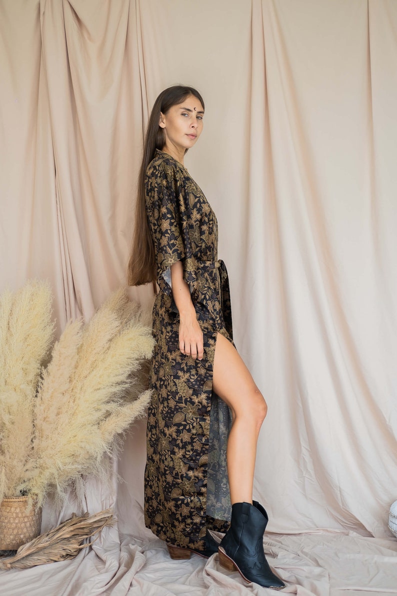 May include: A long, black and gold floral print kimono-style robe with a matching belt. The robe has a high slit on the side and is paired with black ankle boots. The background features neutral-toned fabric and dried ornamental grass.