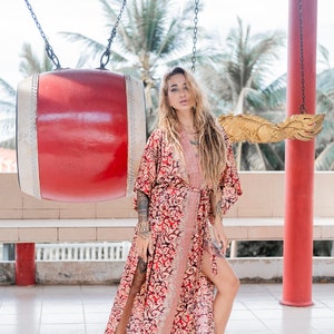 May include: A woman with long blonde dreadlocks wears a red and gold patterned kimono with a long slit up the side. She is standing in front of a large red drum and a golden dragon sculpture.