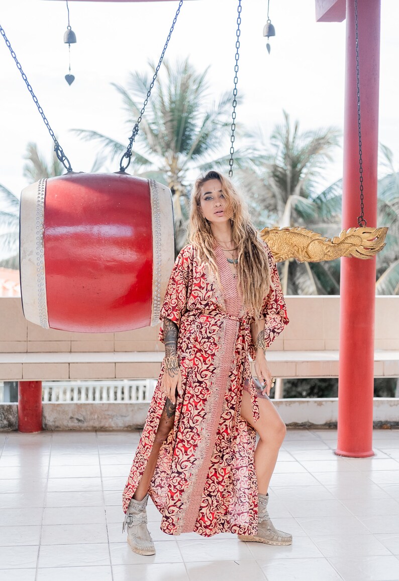 May include: A woman with long blonde dreadlocks wears a red and gold patterned kimono with a long slit up the side. She is standing in front of a large red drum and a golden dragon sculpture.