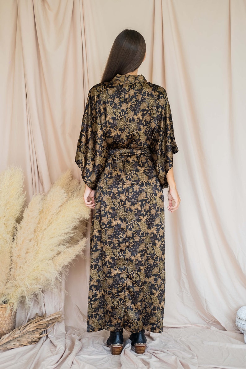 May include: A black and gold floral print kimono-style robe with a matching belt. The long robe features a shawl collar and wide sleeves. The fabric has a shiny finish and a detailed botanical pattern.