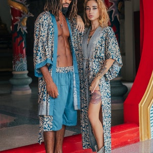 May include: A couple wearing matching blue and gold patterned robes. The man is wearing blue shorts and white sneakers. The woman is wearing brown boots. They are standing in front of a red and gold doorway.
