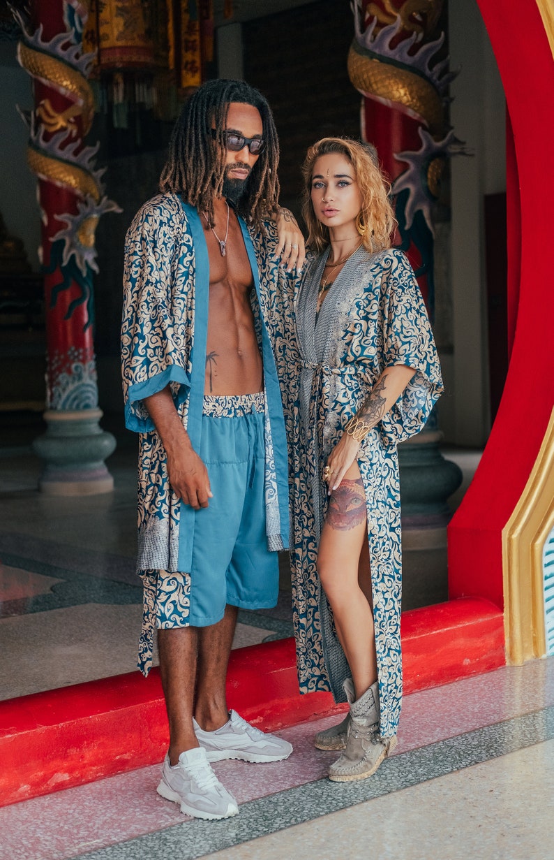 May include: A couple wearing matching blue and gold patterned robes. The man is wearing blue shorts and white sneakers. The woman is wearing brown boots. They are standing in front of a red and gold doorway.