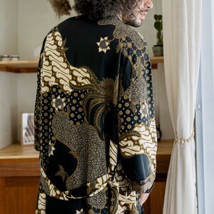 May include: A black robe with a gold and cream batik pattern. The robe features a floral and geometric design. The garment is worn by a person, and the image is taken from behind.