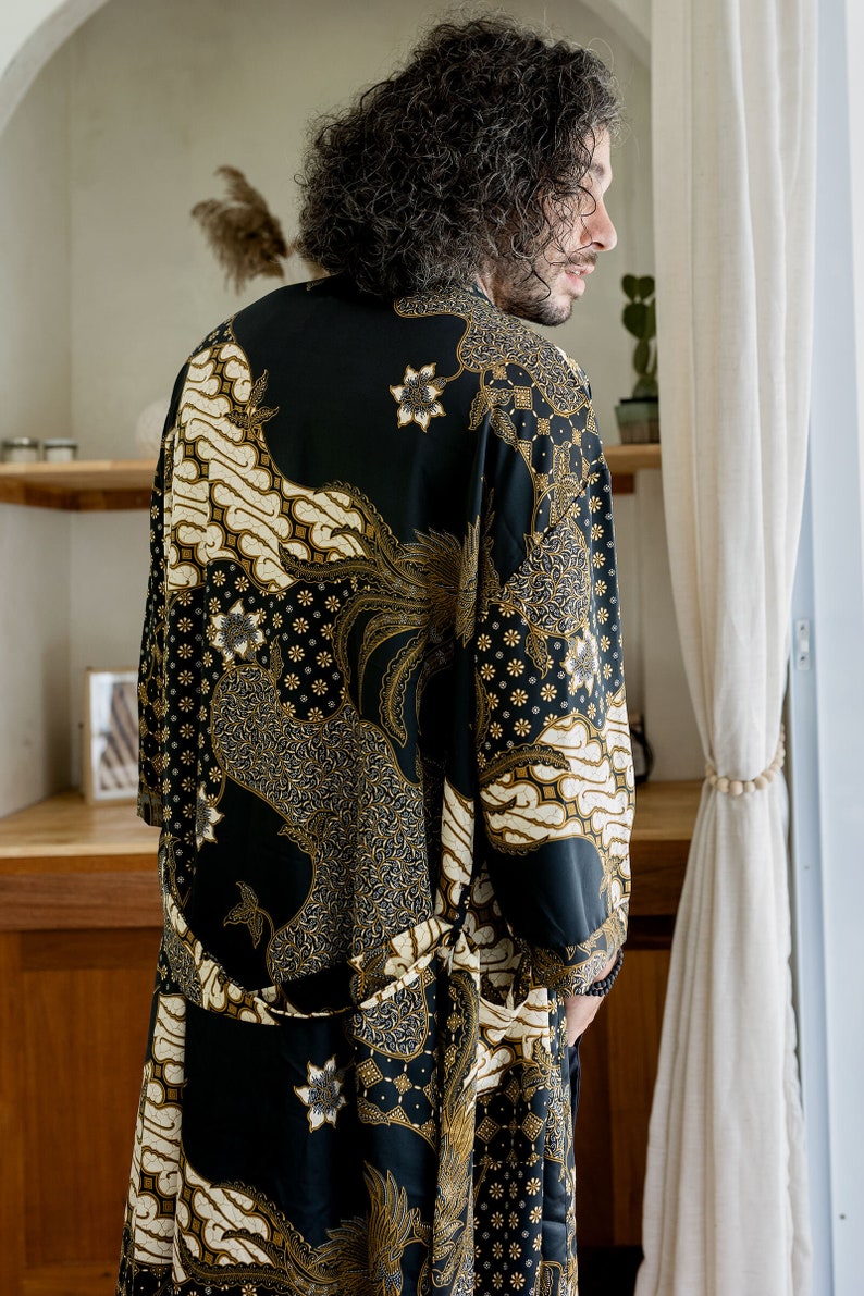 May include: A black robe with a gold and cream batik pattern. The robe features a floral and geometric design. The garment is worn by a person, and the image is taken from behind.