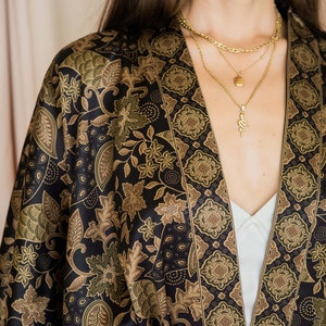 May include: A black kimono-style robe with a floral pattern in shades of brown and green. The robe is open at the front, revealing a white top and gold necklaces. The fabric has a silky sheen.