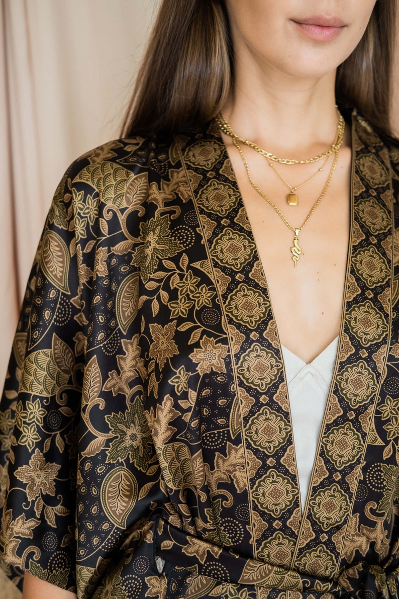 May include: A black kimono-style robe with a floral pattern in shades of brown and green. The robe is open at the front, revealing a white top and gold necklaces. The fabric has a silky sheen.