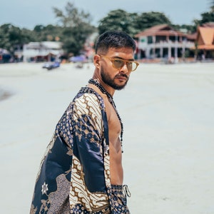 May include: A man wearing a black and gold patterned kimono with a beaded necklace. He is wearing sunglasses and has a beard.
