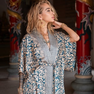 May include: A woman wearing a blue and gold patterned kimono with a gold necklace and earrings. She is standing in front of a red and gold pillar with a dragon design.