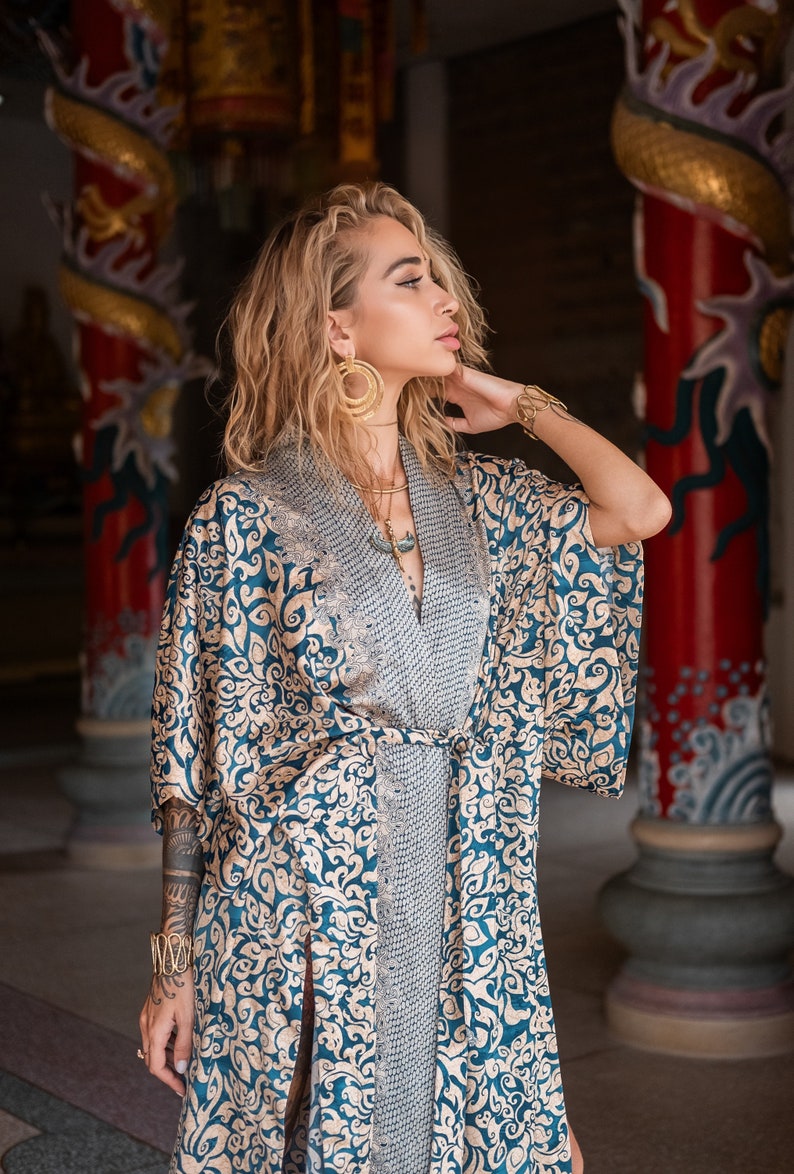 May include: A woman wearing a blue and gold patterned kimono with a gold necklace and earrings. She is standing in front of a red and gold pillar with a dragon design.