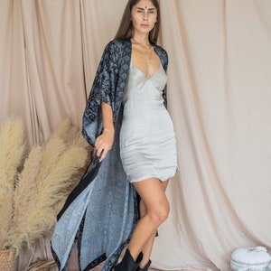 May include: A woman wearing a blue and black patterned kimono over a light grey slip dress. She is wearing black cowboy boots and has a gold necklace on.