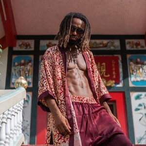 May include: A person wearing a red and gold patterned robe with a burgundy satin shorts. The person is standing on a set of stairs in front of a building with colorful decorations.