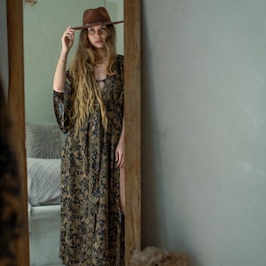 May include: A full-length mirror reflects a woman wearing a brown hat and a long, flowing dress with a floral pattern in shades of black, green, and tan. The dress has a high slit. The mirror is framed in wood, and a woven hat is above it.