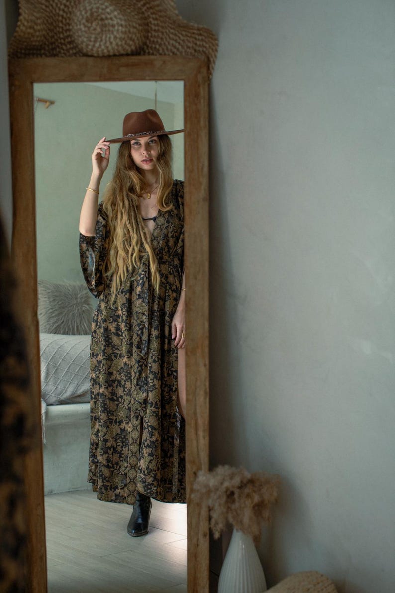 May include: A full-length mirror reflects a woman wearing a brown hat and a long, flowing dress with a floral pattern in shades of black, green, and tan. The dress has a high slit. The mirror is framed in wood, and a woven hat is above it.
