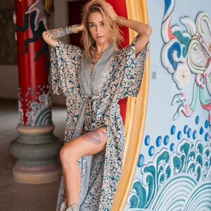May include: A woman wearing a blue and gold patterned kimono with a grey undershirt. She is standing in front of a colorful wall with a dragon design. She is wearing grey suede boots with fringe.