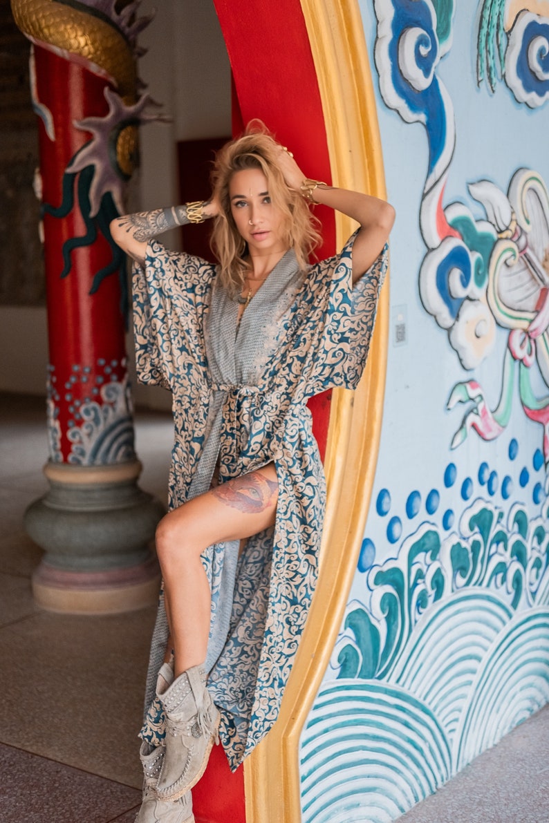 May include: A woman wearing a blue and gold patterned kimono with a grey undershirt. She is standing in front of a colorful wall with a dragon design. She is wearing grey suede boots with fringe.