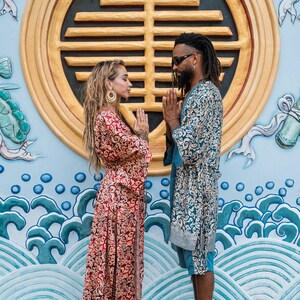 Puede incluir: Dos personas con ropas estampadas se paran frente a una pared azul y dorada con un dise&ntilde;o circular. Ambos se miran con las manos juntas en oraci&oacute;n.