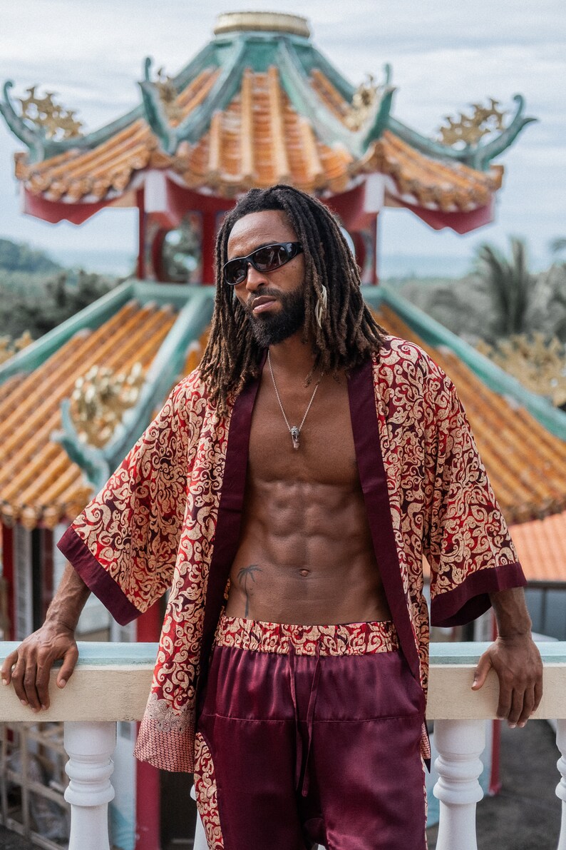 May include: A man wearing a red and gold patterned kimono with a burgundy trim and matching burgundy pants. He is standing in front of a traditional Chinese temple.