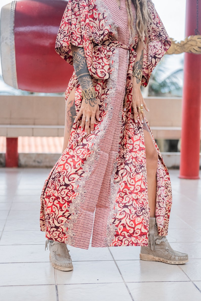 Puede incluir: Una bata de kimono de patr&oacute;n rojo y dorado con una silueta larga y fluida. La bata tiene un lazo de cintur&oacute;n y una abertura lateral. La modelo lleva la bata con un par de botines beige.