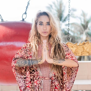 Puede incluir: Una mujer con largas rastas rubias lleva un kimono estampado en rojo y dorado. Est&aacute; de pie frente a un gran gong rojo y tiene las manos juntas en oraci&oacute;n.