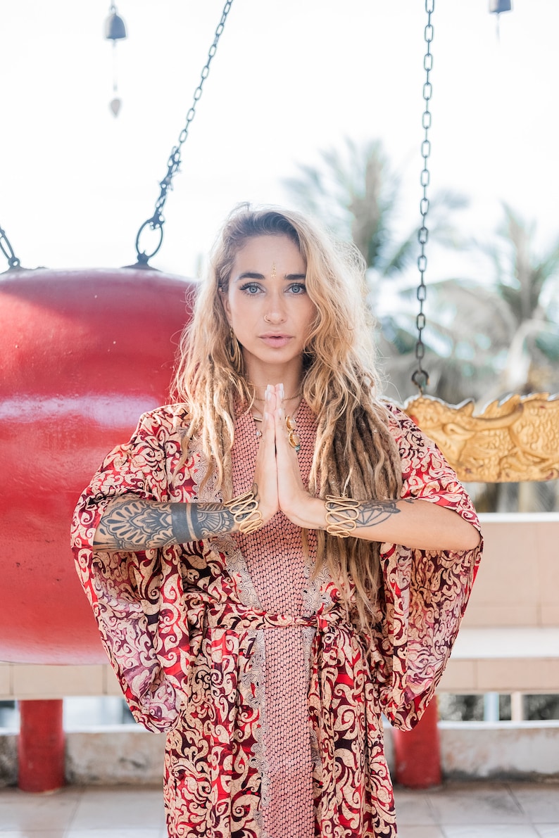 Puede incluir: Una mujer con largas rastas rubias lleva un kimono estampado en rojo y dorado. Est&aacute; de pie frente a un gran gong rojo y tiene las manos juntas en oraci&oacute;n.