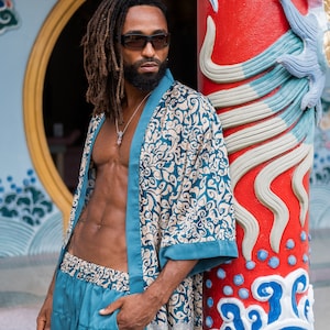 May include: A man with dreadlocks and a beard is wearing a teal blue patterned silk robe and matching pants. He is standing in front of a red pillar with blue and white painted designs.