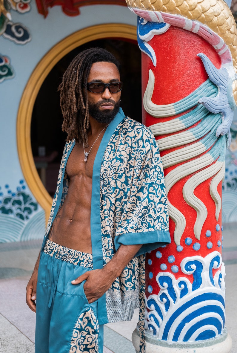 May include: A man with dreadlocks and a beard is wearing a teal blue patterned silk robe and matching pants. He is standing in front of a red pillar with blue and white painted designs.