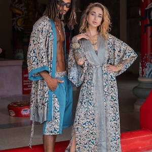 May include: Two people wearing matching blue and gold patterned robes. The woman is wearing a long robe and the man is wearing a shorter robe and matching pants. They are standing in front of a red and gold structure.