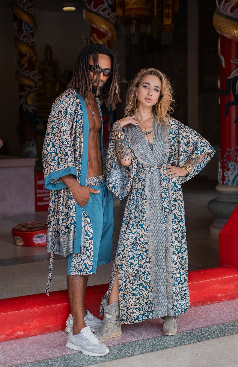 May include: Two people wearing matching blue and gold patterned robes. The woman is wearing a long robe and the man is wearing a shorter robe and matching pants. They are standing in front of a red and gold structure.