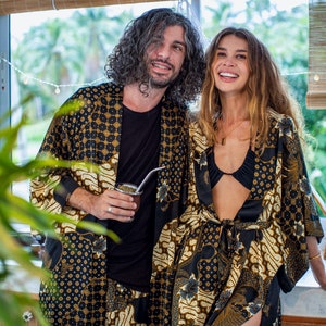 May include: Two people wearing matching black and gold patterned robes. The robes have a batik-style design with floral and geometric elements. One person holds a cup with a metal straw. The background includes a window with a bamboo blind.