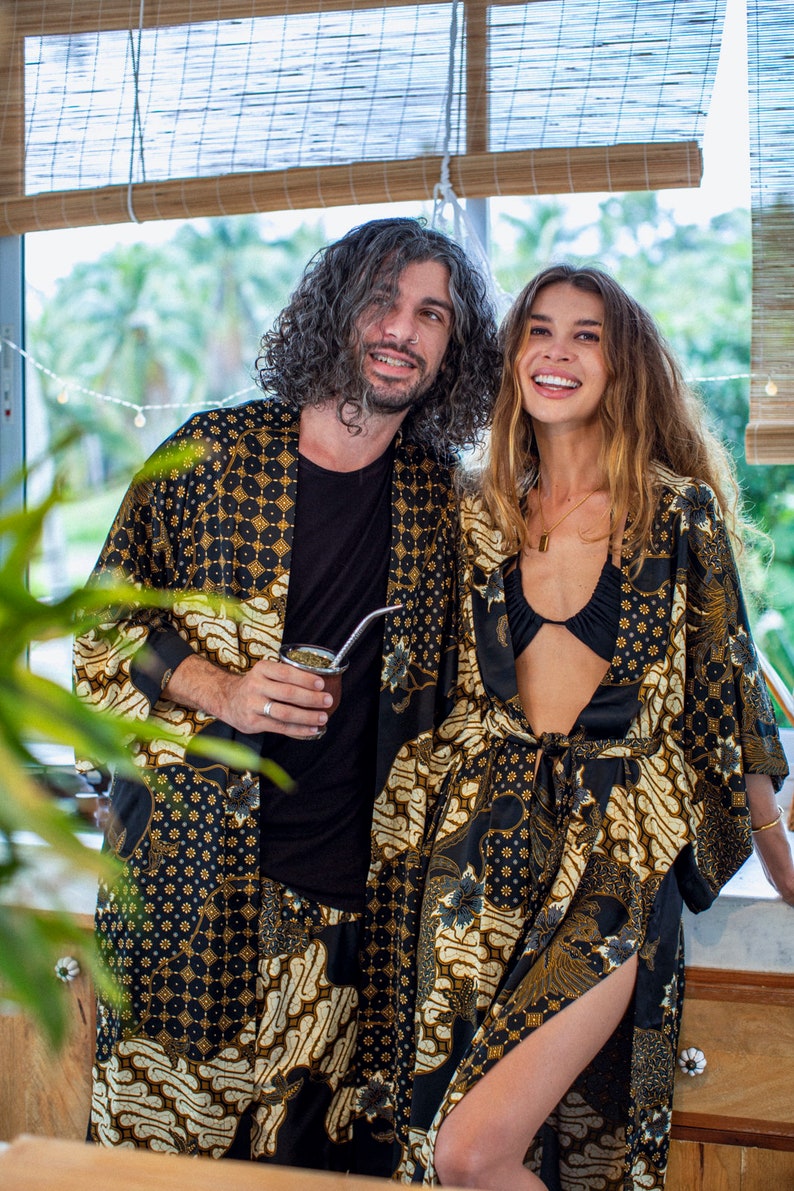May include: Two people wearing matching black and gold patterned robes. The robes have a batik-style design with floral and geometric elements. One person holds a cup with a metal straw. The background includes a window with a bamboo blind.