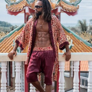 May include: A man wearing a red and gold patterned silk robe with a matching pair of shorts. He is standing on a balcony with a white railing and a traditional Chinese temple in the background.