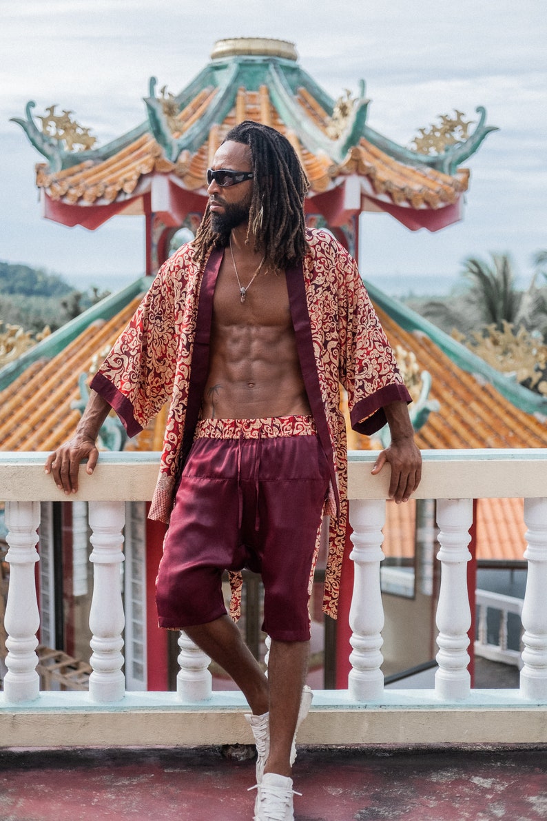 May include: A man wearing a red and gold patterned silk robe with a matching pair of shorts. He is standing on a balcony with a white railing and a traditional Chinese temple in the background.