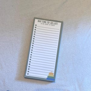 May include: Rectangular notepad with a green border and the text "NAP TIME TO-DO LIST: THE RACE AGAINST THE MONITOR." The notepad has a list of lines for writing and a small stack of colorful shapes at the bottom.