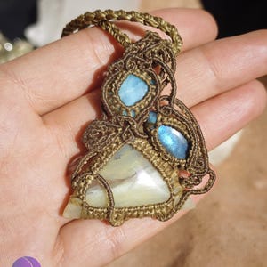 May include: A macrame pendant necklace featuring a large triangular cream-colored stone, a small oval blue stone, and a smaller round blue stone, all encased in intricate brown cord knotting with a braided cord necklace strap.