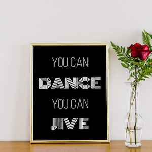 Puede incluir: Una impresión en blanco y negro enmarcada en oro con el texto "You can dance you can jive" sobre una superficie de madera con una sola rosa roja en un jarrón de vidrio transparente.