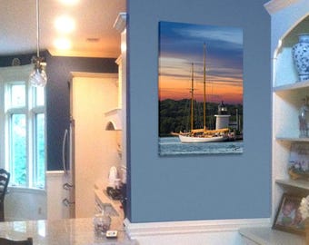 May include: A framed canvas print of a sailboat at sunset. The image features a white sailboat with two masts, a lighthouse, and a colorful sky with shades of orange, blue, and yellow. The print is displayed on a blue wall.