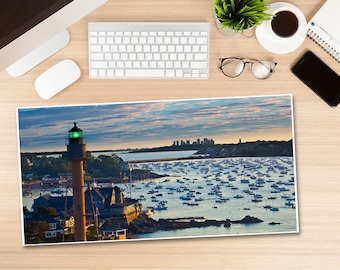 May include: A scenic print of a coastal town at dusk, featuring a lighthouse with a green light, a harbor filled with boats, and a distant cityscape under a cloudy sky. The print is on a wooden desk with a keyboard, mouse, coffee, and a phone.