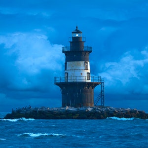 May include: A black and white lighthouse with a light on top, standing on a rocky island in the ocean. The sky is a deep blue with white clouds.