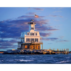 May include: A white lighthouse with a black roof and windows, topped with a small tower and an American flag. The lighthouse is on a rocky island in the ocean, with a wooden pier extending from the shore. The sky is blue with some clouds.