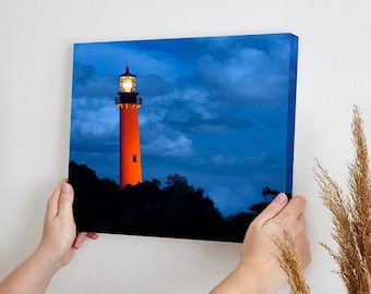 May include: A red and white lighthouse with a light on top, standing tall against a blue sky with clouds. The lighthouse is surrounded by trees and bushes.
