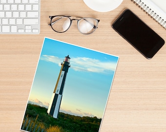 May include: A black and white striped lighthouse with a red light on top, standing tall against a blue sky with white clouds. The lighthouse is surrounded by green grass and a wooden fence.