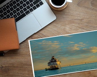 May include: A photograph of a lighthouse on a small island in the middle of the ocean. The sky is blue with white clouds and the sun is setting in the distance. The lighthouse is white with a red roof and a black lantern.